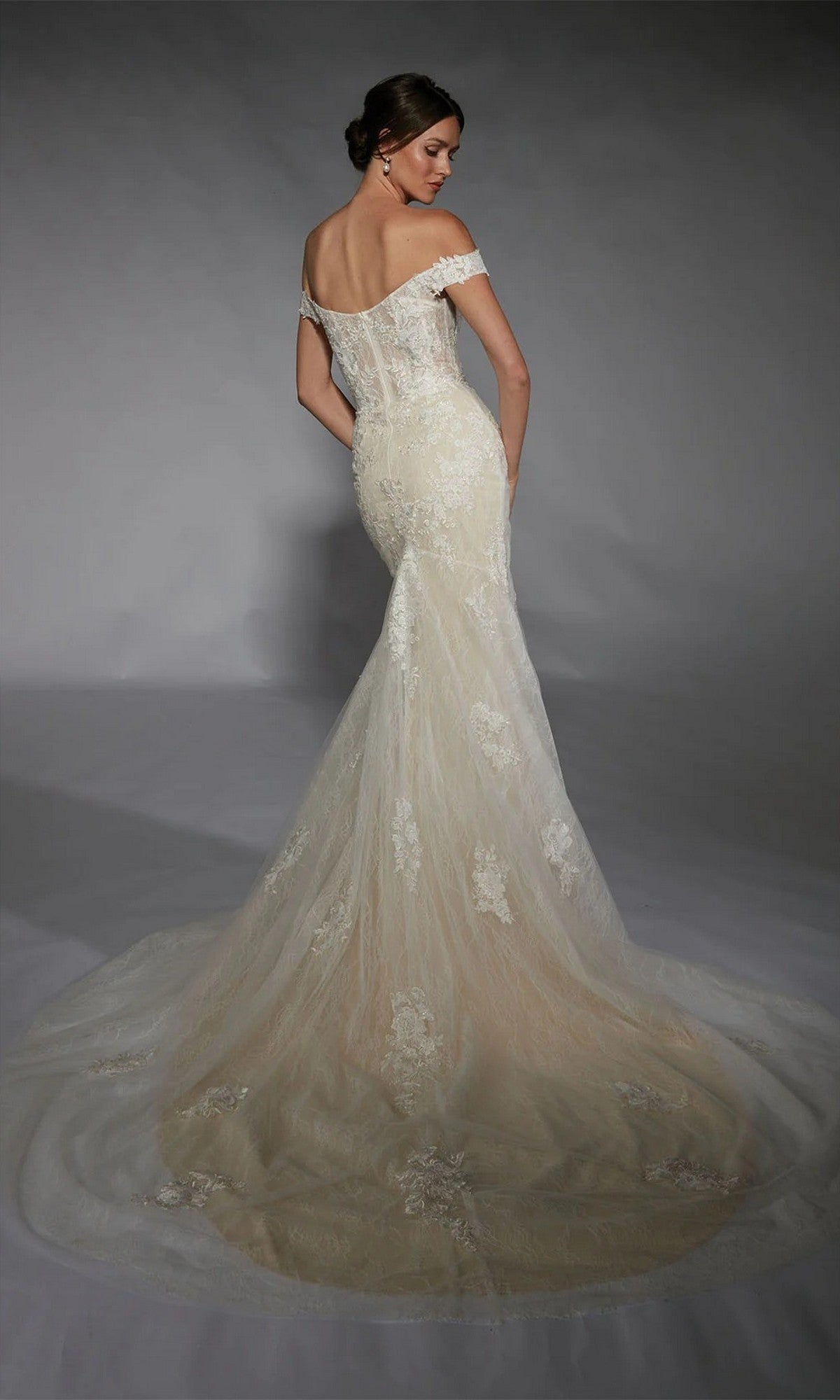 A woman faces away, wearing the Alyce 7118 Long White Dress—an off-the-shoulder lace corset gown with a fitted silhouette and a long, flowing train against a plain gray background.